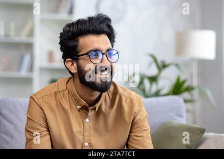Nahaufnahme eines jungen Indianers in Brille, der zu Hause auf der Couch sitzt und mit einem Lächeln zur Seite blickt. Stockfoto
