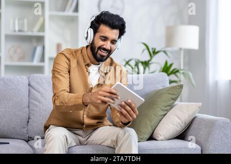 Aufgeregt junger indischer Mann mit Kopfhörern halten digitalen Tablet-Computer, der Online-Videospiele spielt, sitzend auf Couch drinnen lächelnd gutaussehender Mann, der die Freizeit allein zu Hause genießt hochwertiges Foto Stockfoto
