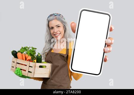 Farmerin, die ein großes Smartphone mit leerem Bildschirm hält, grauer Hintergrund Stockfoto