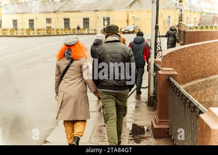 Charmante rothaarige WA charmante rothaarige Frau geht Hand in Hand mit ihrem Freund durch eine stilvolle Herbststadt. Das Paar schlendert die Straße hinunter Stockfoto