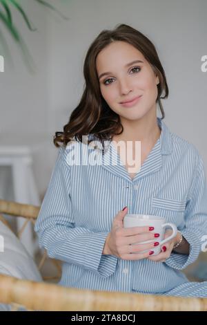 Entspannte junge Frau in gestreiftem Pyjama, die einen Becher hält, ein heiterer Heimagenmorgen mit einem sanften Lächeln und einem frischen Look Stockfoto