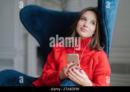 Eine nachdenkliche Frau in einem roten Rollkragen, die Musik auf ihrem Telefon hört, entspannt in einem blauen Stuhl mit hohem Rücken, tagträumt Stockfoto