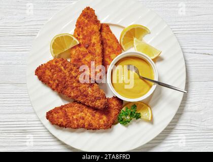 Gebacken im Ofen paniertes Fischfilet mit gelbem Senf und Zitronenscheiben auf weißem Teller auf weißem Holztisch, flach gelegt, Nahaufnahme Stockfoto