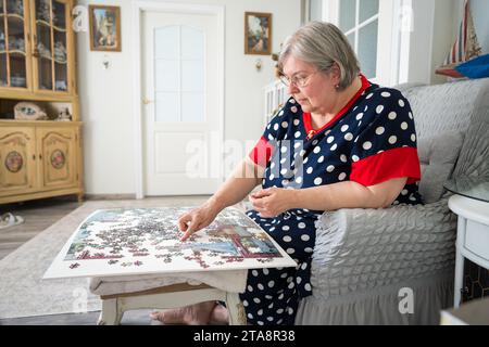 Eine ältere Frau sammelt Puzzle, während sie in einem Stuhl in ihrem hellen Zimmer zu Hause sitzt. Stockfoto