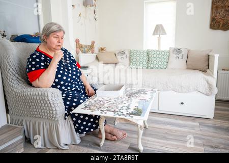 Eine ältere Frau in Gedanken beim Sammeln von Rätseln, während sie auf einem Stuhl in ihrem hellen Zimmer zu Hause sitzt. Stockfoto