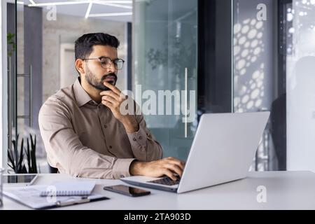 Mann, der mit Laptop im Büro denkt, Geschäftsmann, der Finanzberichte erstellt, auf der Tastatur schreibt, Buchhalter in Brille und Hemd, der Hand bis zum Kinn hält. Stockfoto