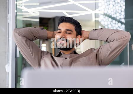 Geschäftsmann ruht Nahaufnahme gut gemacht erfolgreich abgeschlossenes Porträt, Mann mit Händen hinter dem Kopf mit geschlossenen Augen lächelnd, Finanzbuchhalter, der mit Laptop im Büro arbeitet. Stockfoto