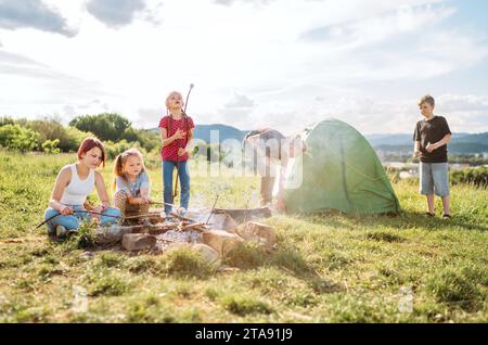 Drei Schwestern lachen fröhlich und rösten auf Stöcken über der Lagerfeuerflamme Sümpfe und Bonbons, während zwei Brüder das grüne Zelt aufschlagen. Glücklich Stockfoto