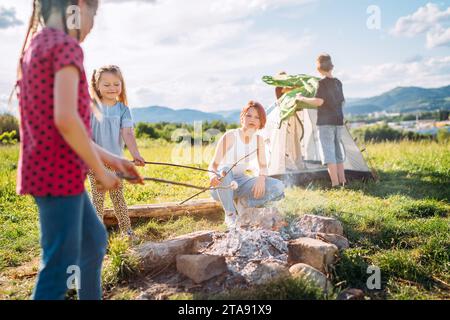 Drei Schwestern machen Picknick, rösten Marshmallows und Süßigkeiten auf Stöcken über Lagerfeuer, während zwei Brüder das grüne Zelt aufbauten. Glückliche Familie, Outd Stockfoto