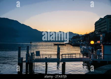 Atemberaubende Aussicht auf den abendlichen Sonnenuntergang auf den Comer See und einige Boote ankern und Gebäude an der Küste (Hotel?), Pier und elektrisches Licht. Kurze Reise während Weihnachten wir Stockfoto