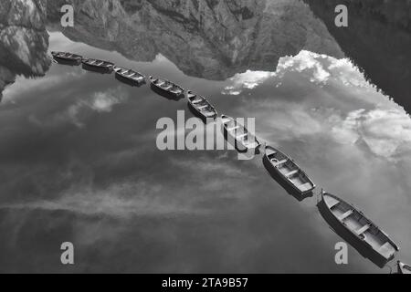 Einfarbiger, künstlerischer, rechtwinkliger Blick auf Holzboote in Reihe zwischen spiegelnden Gipfeln und Himmel auf einem Pragser See in den Dolomiten. Stockfoto