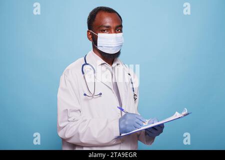 Porträt eines schwarzen Mannes, der eine Gesichtsmaske und einen Labormantel trägt, während er medizinische Forschungsdokumente hält. Männlicher Gesundheitsspezialist posiert für die Kamera, während er einen Stift und ein Klemmbrett greift. Stockfoto