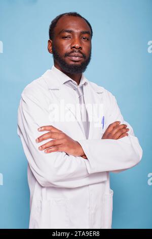 Afroamerikaner, männlicher Arzt, der vor einem isolierten blauen Hintergrund steht. Ernster schwarzer Mann, der einen weißen Labormantel trägt, posiert mit überkreuzten Armen vor der Kamera. Stockfoto