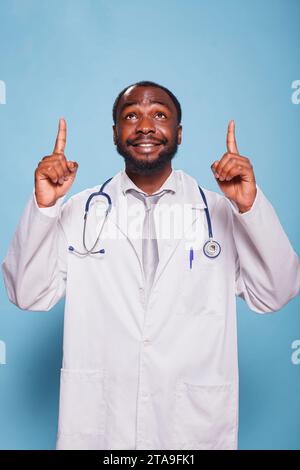 Vor isoliertem Hintergrund zeigt der lächelnde Arzt beide Zeigefinger nach oben, während er Labormantel und Stethoskop trägt. Optimistischer Arzt, der eine hoffnungsvolle Handgeste zum Himmel macht. Stockfoto