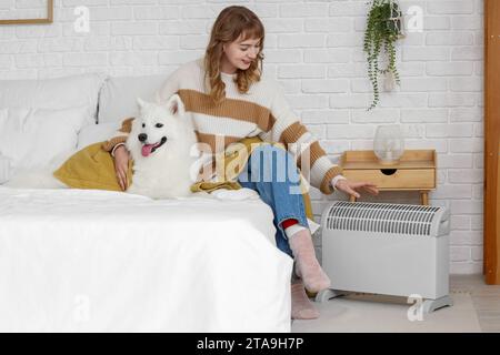 Junge Frau mit Decke und Samoiden Hund wärmt sich nahe dem Heizkörper im Schlafzimmer Stockfoto