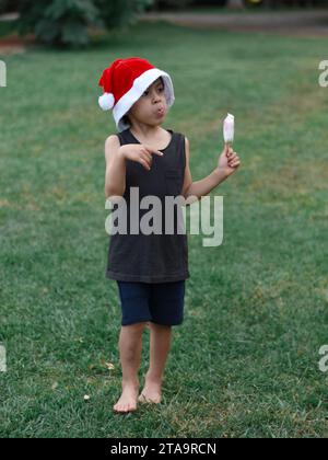 Entspannter Latin Boy, der im Sommer ein Eis genießt und Weihnachten vorwegnimmt, trägt Shorts, ein Tanktop und einen Weihnachtsmann-Hut auf dem Rasen seines Gartens. Stockfoto