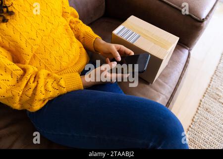 Mittelteil einer birassischen Frau in gelbem Pullover mit Paket mit Smartphone zu Hause Stockfoto