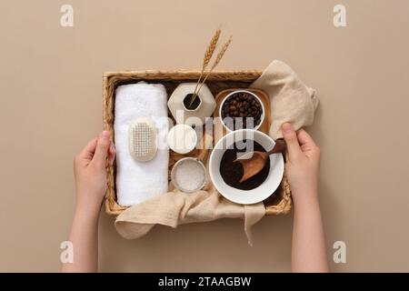 Zwei Hände einer Frau, die einen Korb mit Badegegenständen, Kaffeebohnen und Kaffeepulver, Badesalz, Kerze und Handtuch auf hellbraunem Hintergrund hält Stockfoto