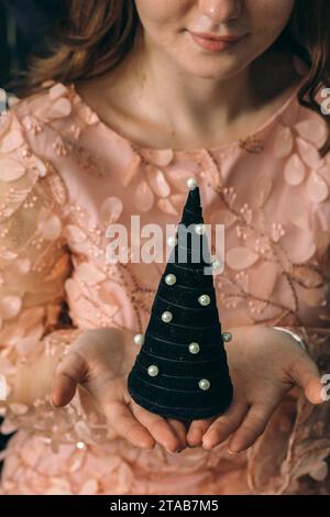 Nahaufnahme von weiblichen Händen, die einen kleinen schwarzen Weihnachtsbaum halten, feiern und Weihnachtsfeiertage genießen. Zartes Rosa Stockfoto