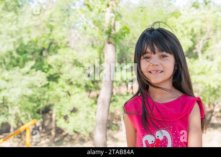 Wunderschönes lateinisches Kind, das für ein Foto lächelt, in einem Park voller Bäume und Grüntöne. Stockfoto