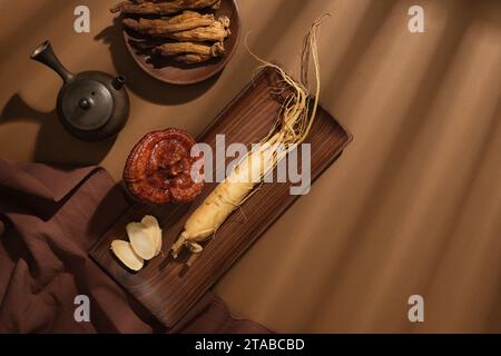 Draufsicht auf Ginseng-Wurzel und Lingzhi-Pilz auf Holztablett mit Teekanne und Tuch auf braunem Hintergrund. Sammlung von Kräutern, um eine Tonne vorzubereiten Stockfoto