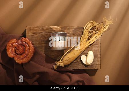 Auf einem hölzernen Podium, ein Kosmetikglas mit Wurzel und Scheiben Ginseng, Lingzhi-Pilz und braunem Tuch auf braunem Hintergrund. Konzept für Cosmet Stockfoto