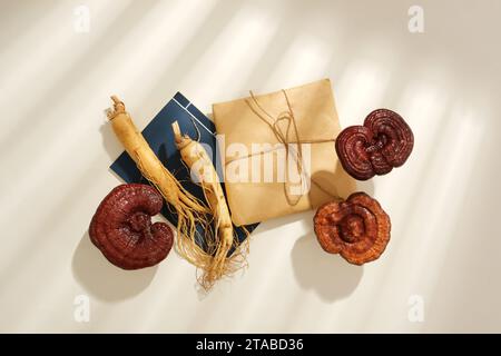 Blick von oben auf Lingzhi-Pilze und Ginseng-Wurzeln, verziert mit Medikamentenverpackung und blauem Notizbuch auf weißem Hintergrund mit Schatten des Fensters. Minimal Stockfoto