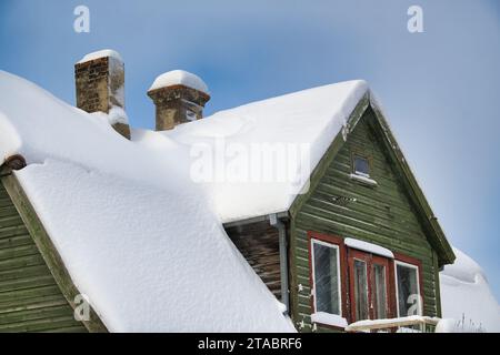 Eine ruhige Winterszene, die sich auf ein grünes Holzhaus konzentriert. Das Dach des Hauses ist stark mit weißem Schnee bedeckt, wobei zwei Schornsteine aus dem dicken Lila ragen Stockfoto