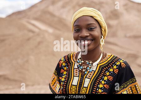 Porträt einer jungen Schwarzen Frau, die ein Outfit mit traditionellen afrikanischen Ornamenten trägt, während sie in der Wüste posiert und lächelnd im Kopierraum erscheint Stockfoto