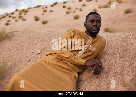 Porträt eines afroamerikanischen Mannes in langem Kaftan-Kleid, der auf Sanddünen in der Wüste liegt und wehmütig wegblickt Stockfoto