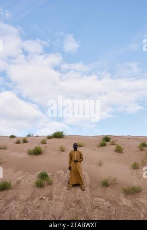 Vertikale Weitwinkelaufnahme des Schwarzen Mannes, der auf einer Sanddüne in der Wüste steht und traditionellen Kaftan trägt, Kopierraum Stockfoto