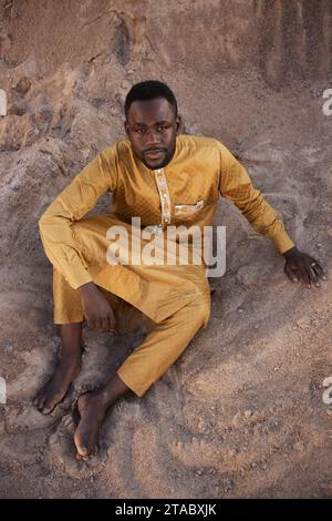 Hochwinkelporträt eines schwarzen jungen Mannes, der traditionellen Kaftan trägt und im Schatten auf Sand sitzt und in die Kamera blickt Stockfoto