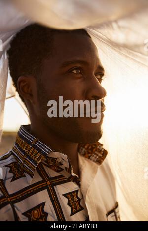 Modeporträt eines lächelnden Schwarzen Mannes mit traditionellem Muster, der draußen mit Schleier bedeckt schaut Stockfoto