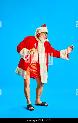 Foto in voller Länge eines glücklichen, fröhlichen älteren Mannes in Gläsern, der Spaß hat, mit Cocktail in der Hand vor blauem Studiohintergrund tanzt. Stockfoto