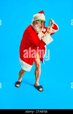 Porträt in voller Länge fröhlich, älterer Mann, der mit einer alten Kamera vor blauem Studiohintergrund fotografiert. Holly Jolly Abenteuer. Stockfoto