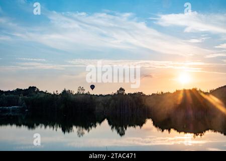 Dieses Bild wurde in dem Moment aufgenommen, in dem die Dämmerung aufbricht, und zeigt einen ruhigen See, der den erwachenden Himmel reflektiert. Die Sonne ragt über den Horizont, streut strahlende Strahlen und erweckt die Welt mit sanftem Licht. Ein einsamer Heißluftballon schwimmt sanft in der Ferne und verleiht der morgendlichen Ruhe einen Hauch von Abenteuer und Laune. Diese ruhige Szene ist eine harmonische Mischung aus menschlichem Anspruch und natürlicher Eleganz. Sonnenaufgang im Heißluftballon über einem See mit Spiegeln. Hochwertige Fotos Stockfoto