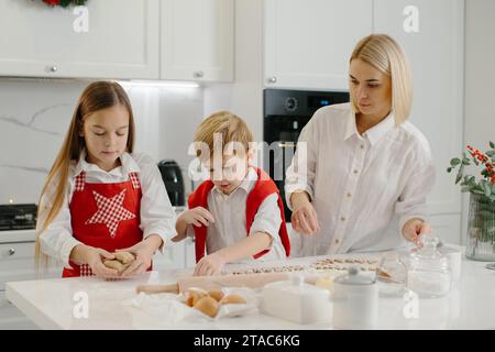 Eine Mutter bringt ihrer Tochter und ihrem kleinen Sohn bei, wie man Kekse macht. Eine Frau und ihre Kinder bereiten Weihnachtskekse mit einer Teigform zu. Stockfoto