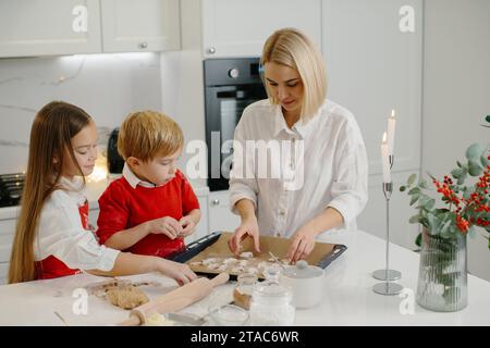 Junge, glückliche Mutter mit Kindern, die Weihnachtskuchenkekse machen und sie auf ein Tablett in der Küche stellen. Stockfoto