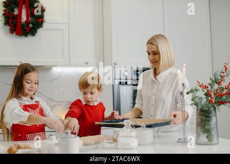 Junge, glückliche Mutter mit Kindern, die Weihnachtskuchenkekse machen und sie auf ein Tablett in der Küche stellen. Stockfoto