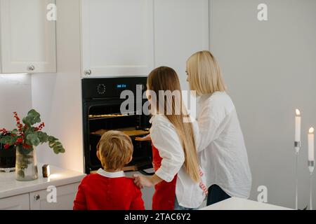 Mutter und Kinder backen Weihnachtskuchen in einer modernen Küche. Mom stellt ein Tablett mit Keksen in den Ofen. Vorbereitung eines Familienurlaubs Stockfoto