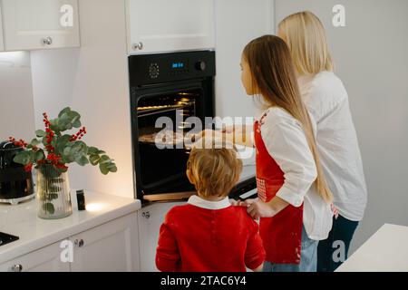 Mutter und Kinder backen Weihnachtskuchen in einer modernen Küche. Mom stellt ein Tablett mit Keksen in den Ofen. Vorbereitung eines Familienurlaubs Stockfoto