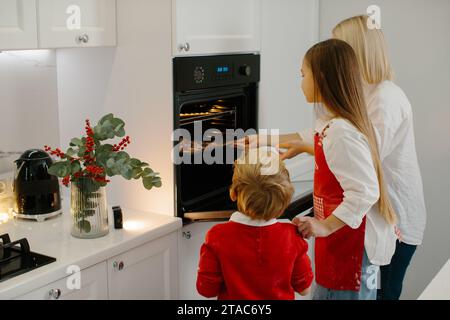 Mutter und Kinder backen Weihnachtskuchen in einer modernen Küche. Mom stellt ein Tablett mit Keksen in den Ofen. Vorbereitung eines Familienurlaubs Stockfoto