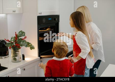 Mutter und Kinder backen Weihnachtskuchen in einer modernen Küche. Mom stellt ein Tablett mit Keksen in den Ofen. Vorbereitung eines Familienurlaubs Stockfoto