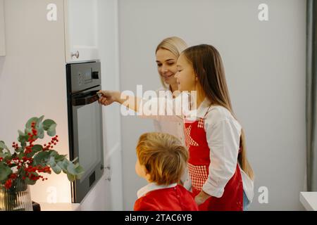 Mutter und Kinder backen Weihnachtskuchen in einer modernen Küche. Mom stellt ein Tablett mit Keksen in den Ofen. Vorbereitung eines Familienurlaubs Stockfoto