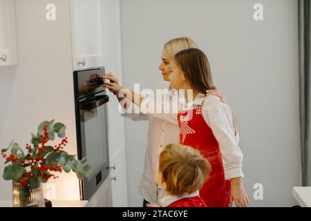 Mutter und Kinder backen Weihnachtskuchen in einer modernen Küche. Mom stellt ein Tablett mit Keksen in den Ofen. Vorbereitung eines Familienurlaubs Stockfoto