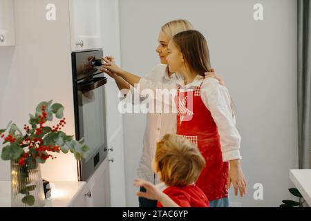 Mutter und Kinder backen Weihnachtskuchen in einer modernen Küche. Mom stellt ein Tablett mit Keksen in den Ofen. Vorbereitung eines Familienurlaubs Stockfoto