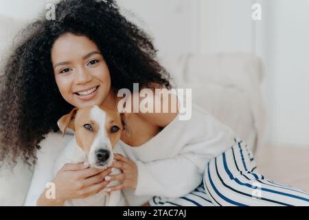 Frau mit lockigen Haaren in einem gemütlichen Pullover, der ihren Hund umarmt, beide sehen zufrieden aus. Stockfoto