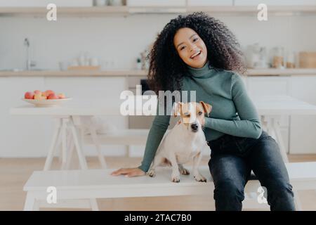 Lächelnde Frau im Rollkragen mit ihrem Hund an einem Küchentisch, strahlend Freude. Stockfoto
