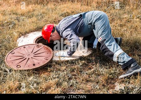 Klempner in einem Schutzhelm beugte sich über einen Wasserbrunnen, um den Wasserzähler zu überprüfen. Inspektion und Reparatur von Leitungen. Stockfoto