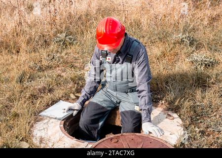 Ein Klempner in Overall und Helm sitzt auf der Seite einer Brunnenluke mit Dokumenten und überprüft den Wasserzähler. Stockfoto